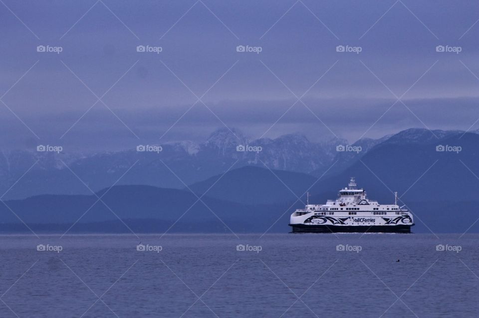 A stark white ferry crossing the ocean strait on calm blue waters. The rolling hills on the coast and the snow capped mountains provide a stunningly beautiful backdrop. 
