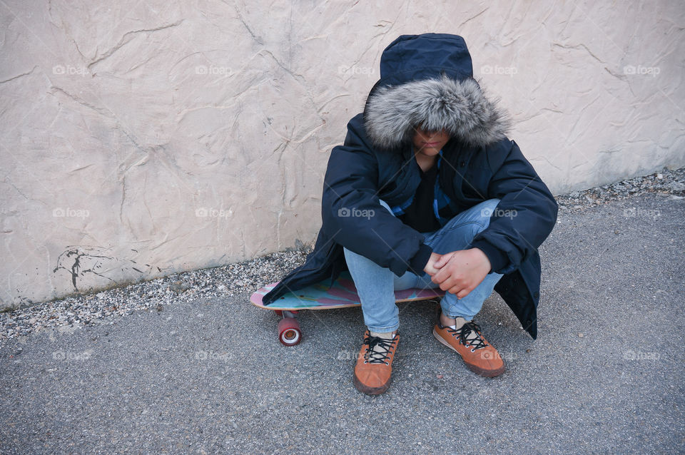 Boy wearing winter jacket sitting on skateboard next to concrete wall