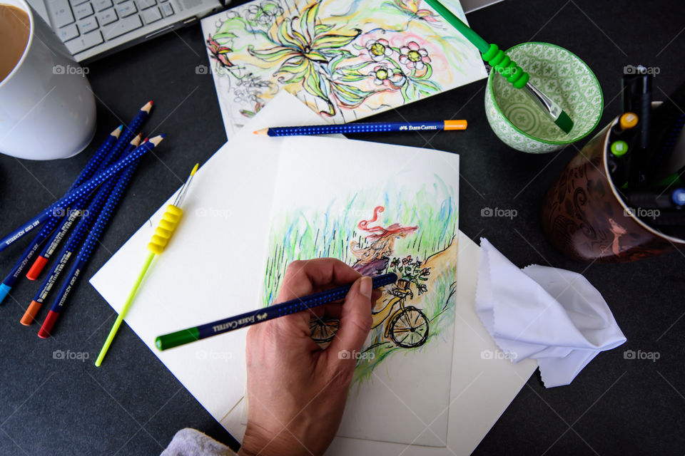 Flat lay of hand holding Faber-Castell Aquarelle Colored watercolor pencils sketching girl on bicycle in countryside with sketchbook on table with laptop and coffee conceptual work life balance, creativity, hobby artist lifestyle photography 