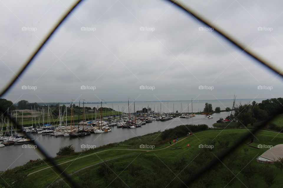 Through a wire fence looking at a harbor in Europe in the summer time with green grass.