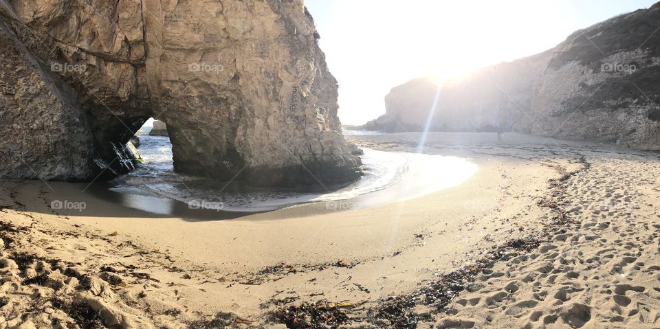 A panoramic view of sun rays kissing the rocky cliffs of shark fin cove as the waves crash through a hole in the rock. 