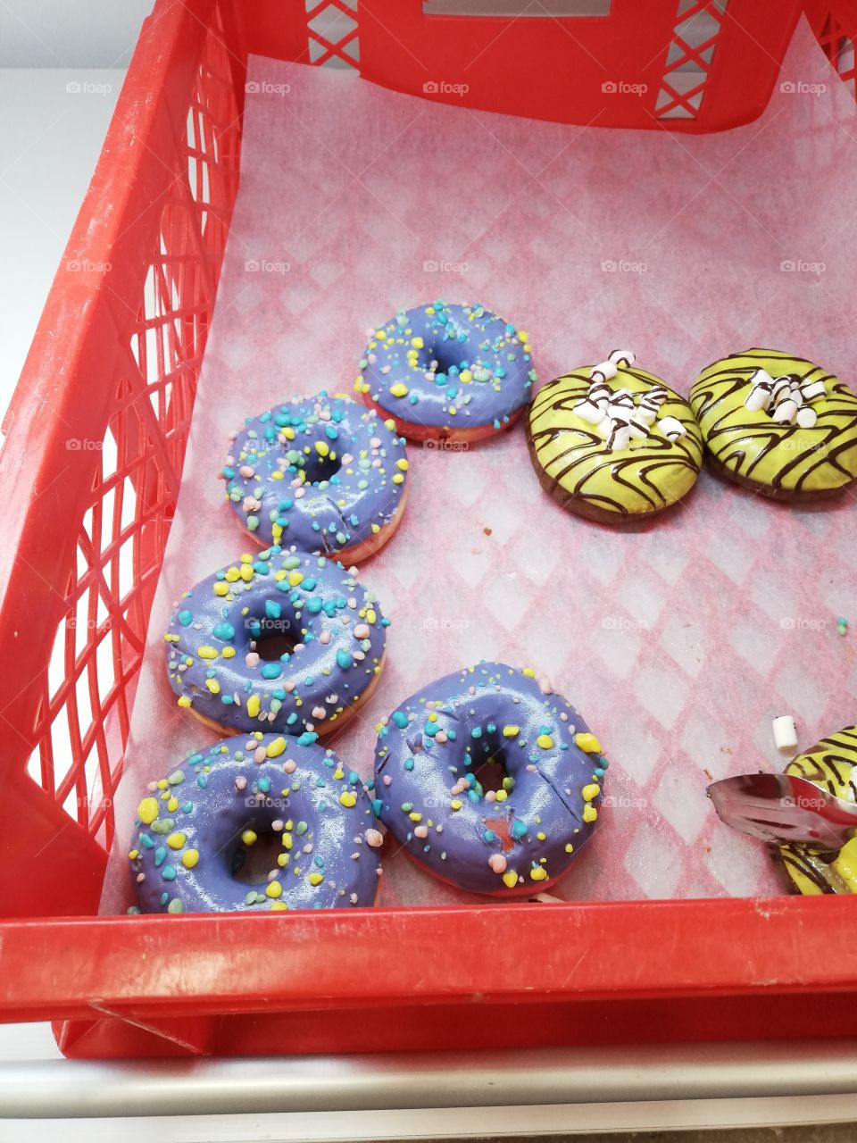 Some yellow and blue donuts with sugary coating, marshmallow, nonpareils and chocolate stripes are in a red plastic box. The see-through baking paper is too big to cover almost empty bottom.