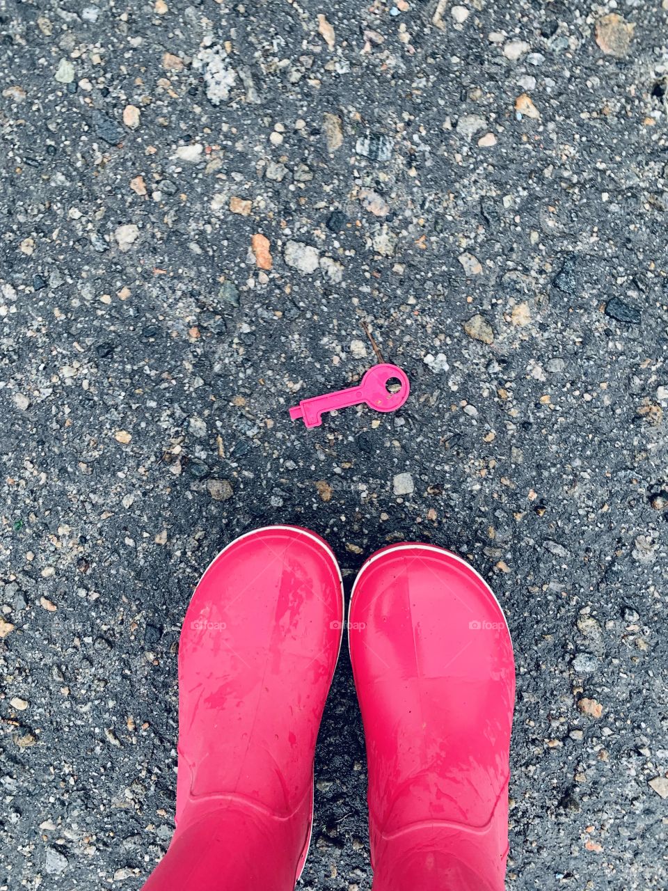Find a clue! Colorful world. Pink rain boots and toy key at wet asphalt 