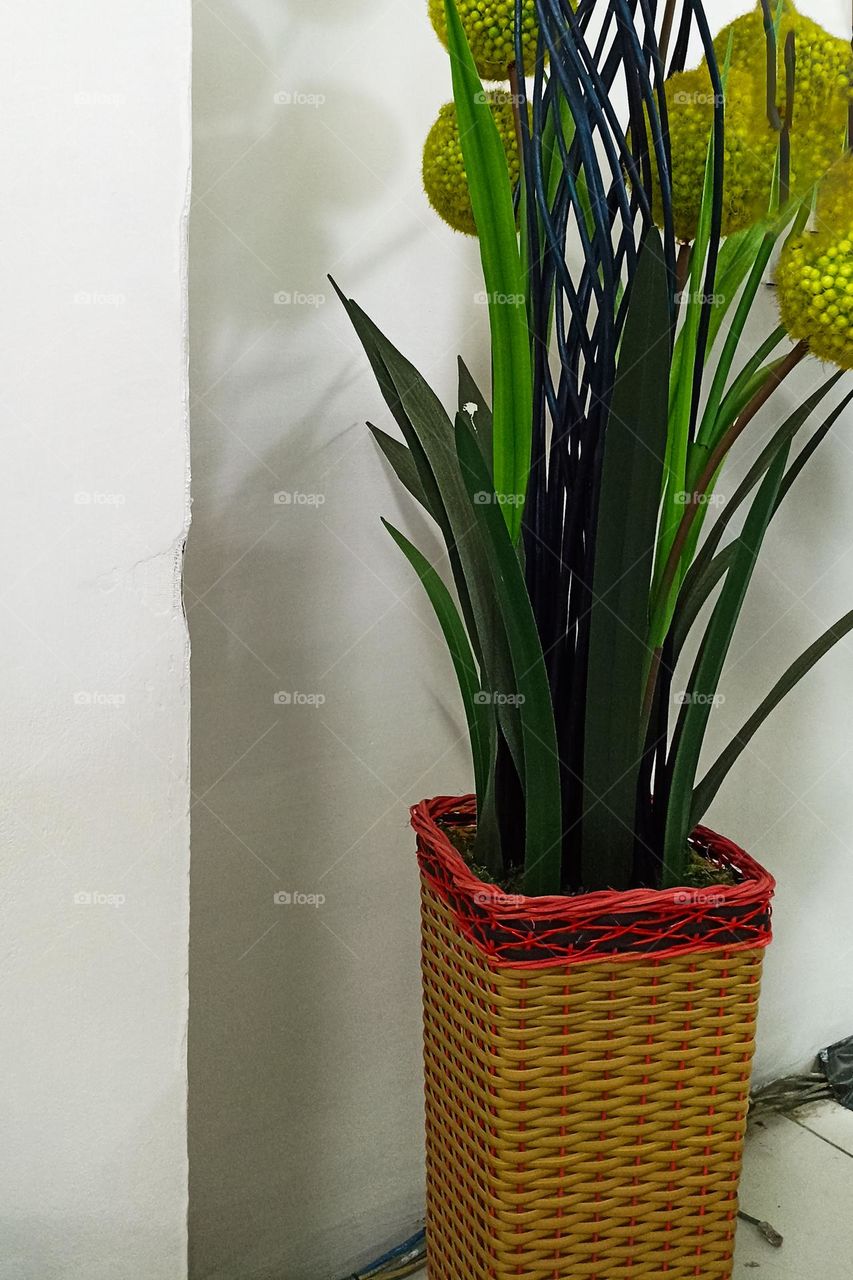 a flower vase made of wicker, with green plants and yellow flowers inside, placed near a white wall