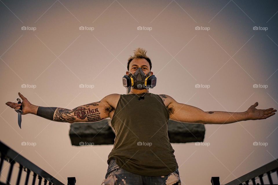 Punk man with gas mask, knife and tattoo, standing in front of a cross