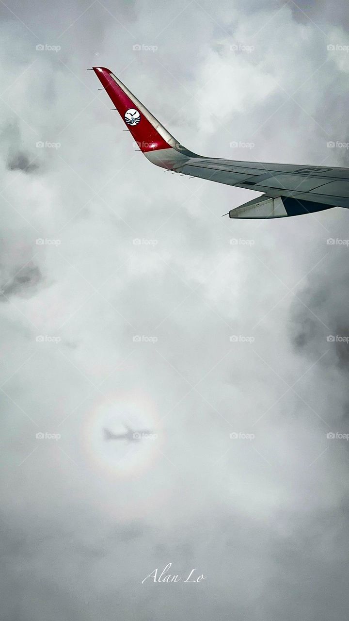 An airplane wing is set against a cloudy sky. Below the wing, there is a circular optical effect known as a "glory," showing the shadow of the same airplane.