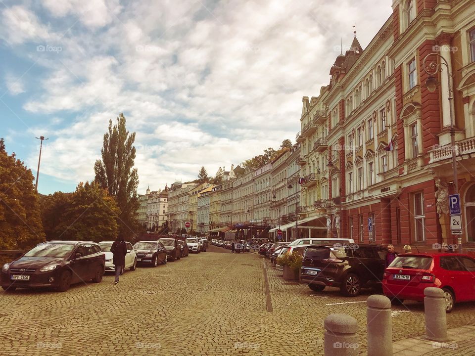 A series of buildings on the street, the street surface is paved with stones, 2 sides have many cars parked , In the sky a lot of clouds . Some people are walking