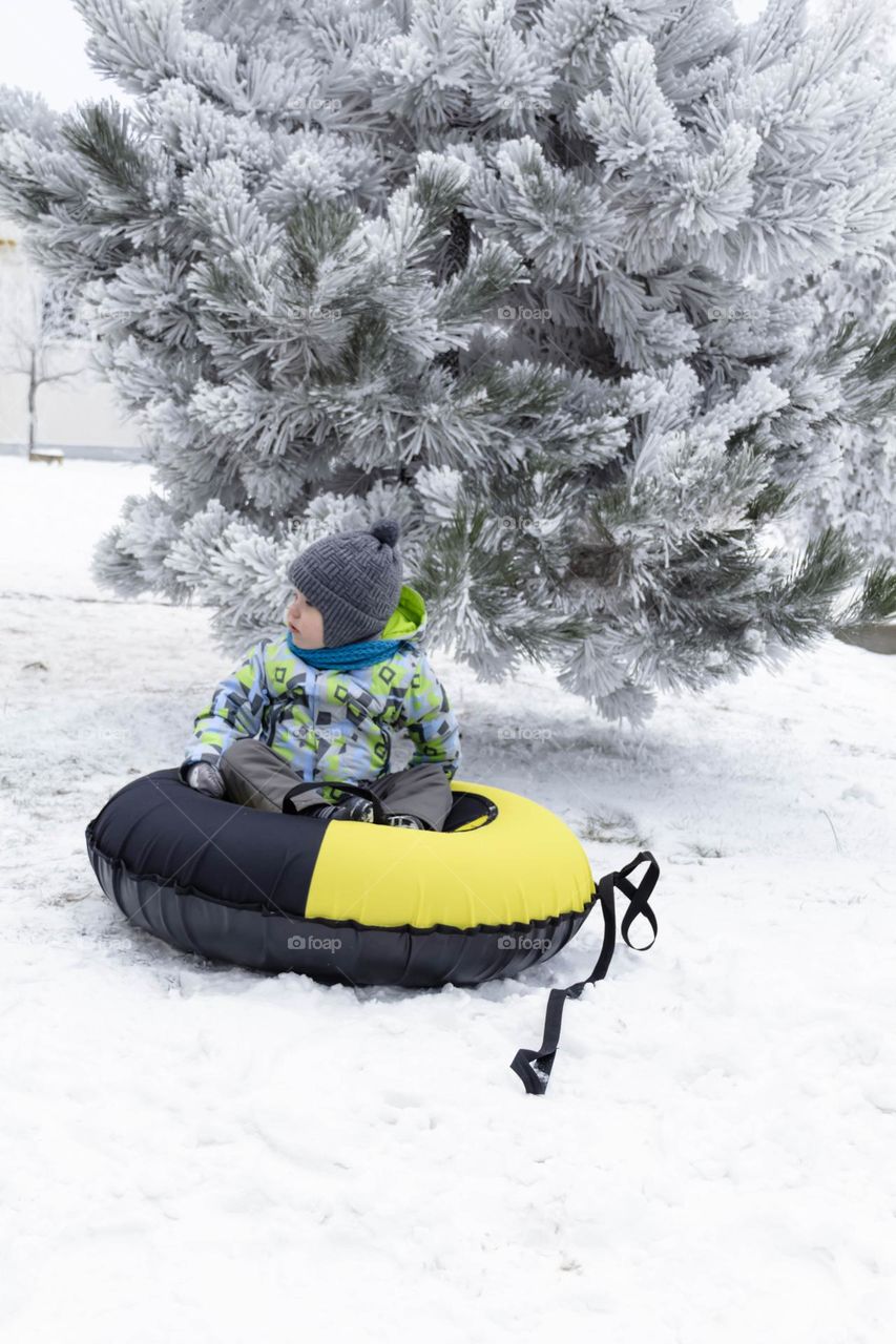 A small, carefree boy walks in the white snow in winter and rides a tubing in the park, near trees in the snow.