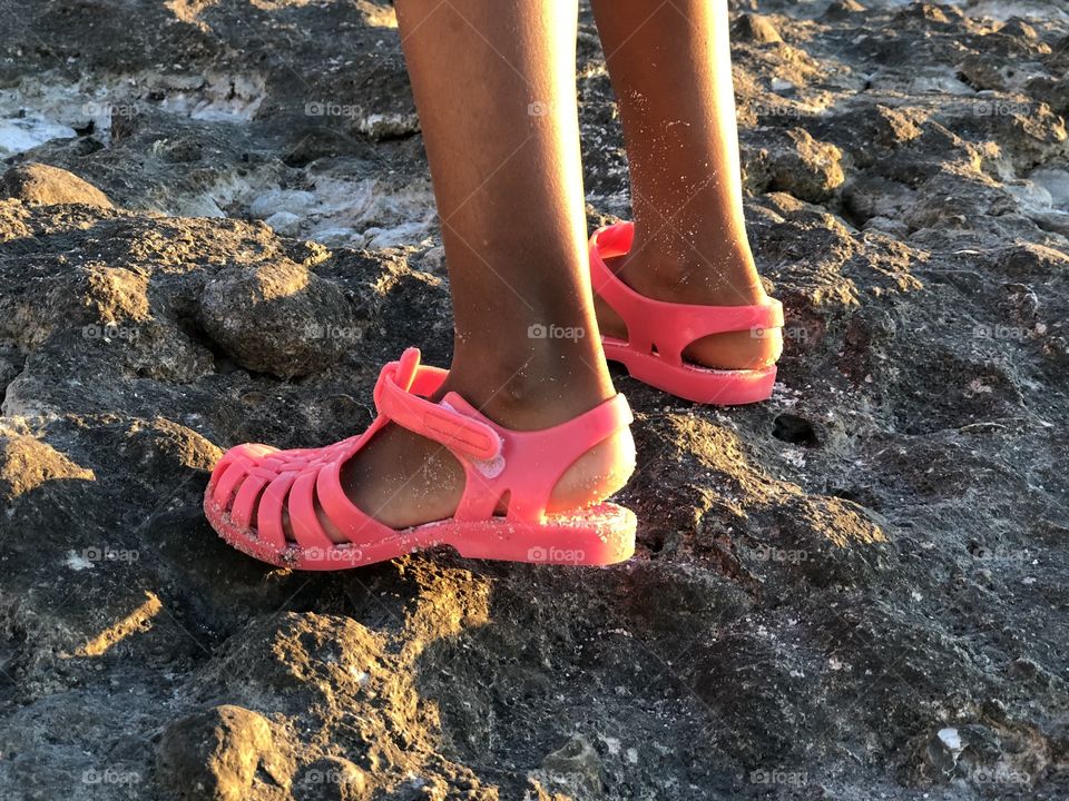 A girl standing on a rock at a beach with sandy shoes