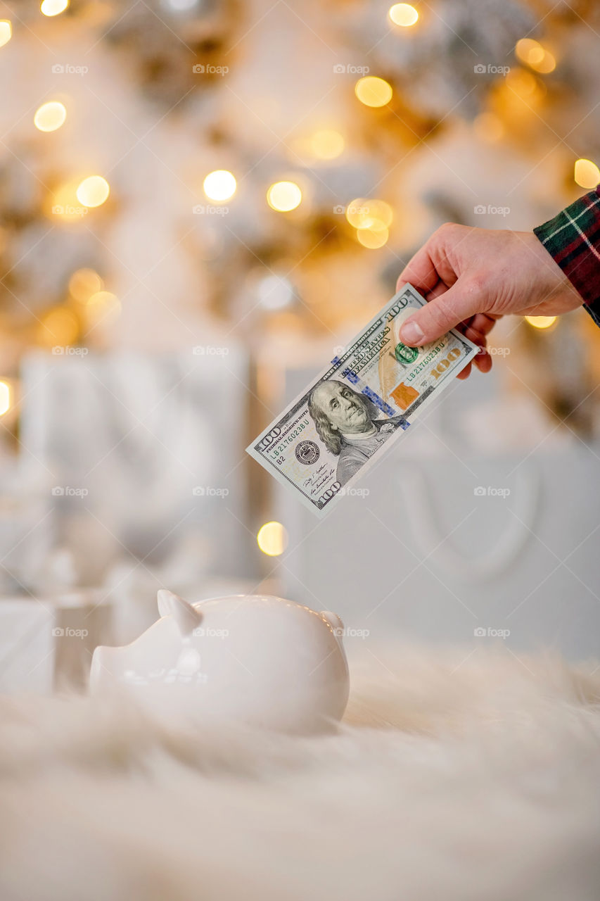 piggy bank with 100 dollars banknote in festive New Year atmosphere of scenery.  Magical bokeh with Christmas tree and bright lights.