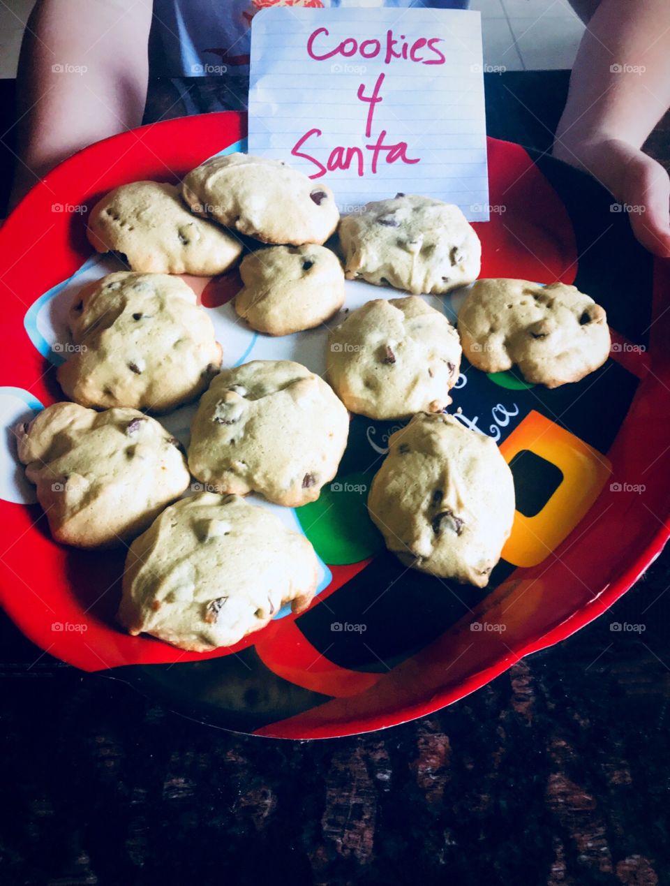 A child holding a plate of chocolate chip Cookies for Santa on a red Santa Clause plate on Christmas Eve. 