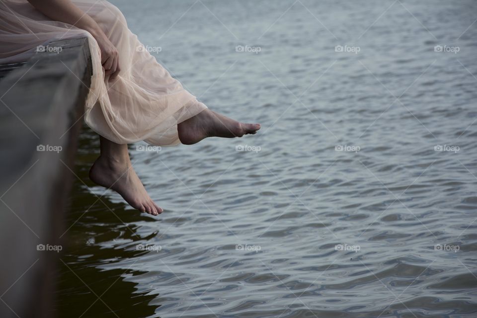 feet above water. woman barefoot sitting in pretty dress on the deck above lake