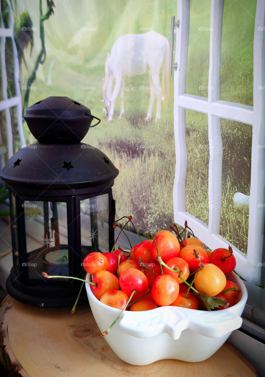 On the windowsill is a white cup in the shape of a heart, in which lies a yellow-pink cherry.Nearby is a black lantern. Outside the window is a green meadow with a white horse. There is a grid on the window. Summer landscape with still life