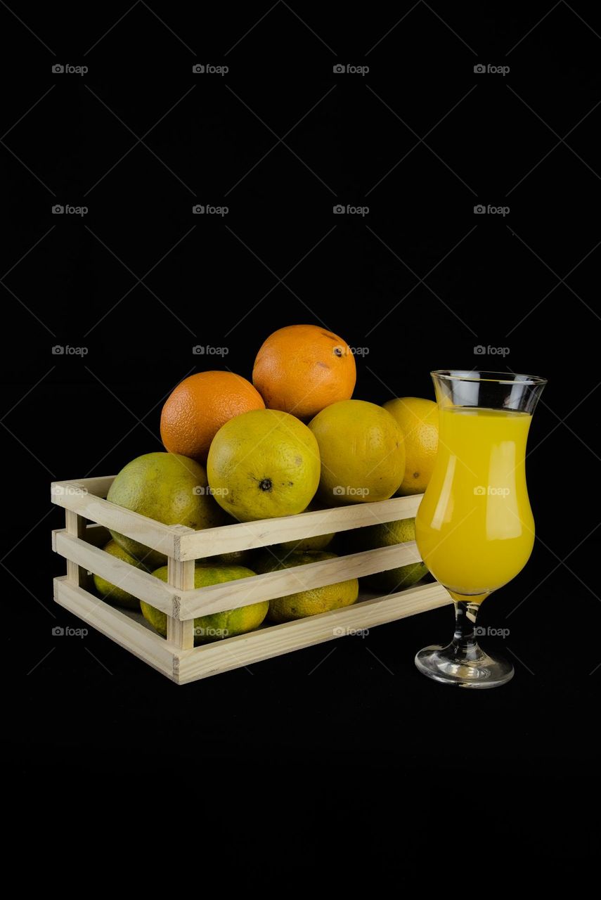 Wooden box with oranges and crystal glass with juice on black background