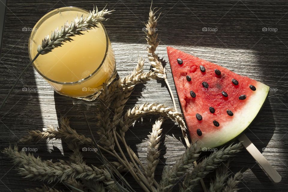 A piece of ripe, red watermelon and cold juice, a cocktail with ice and ears of wheat stand on a wooden table.  Summer drinks and snacks.