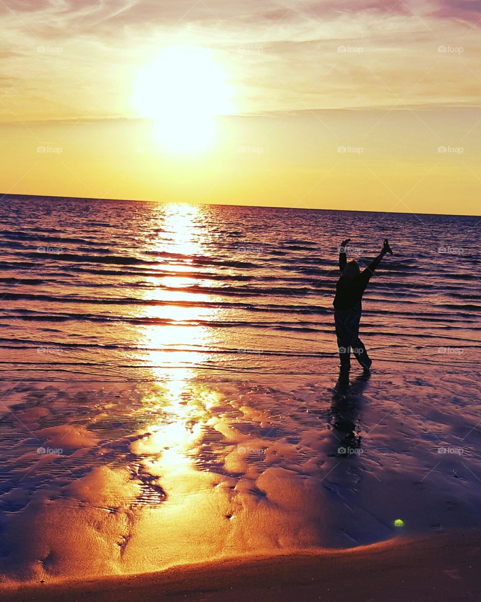 A lone figure walks the beach at sunset and enthusiasticly jumps for joy.