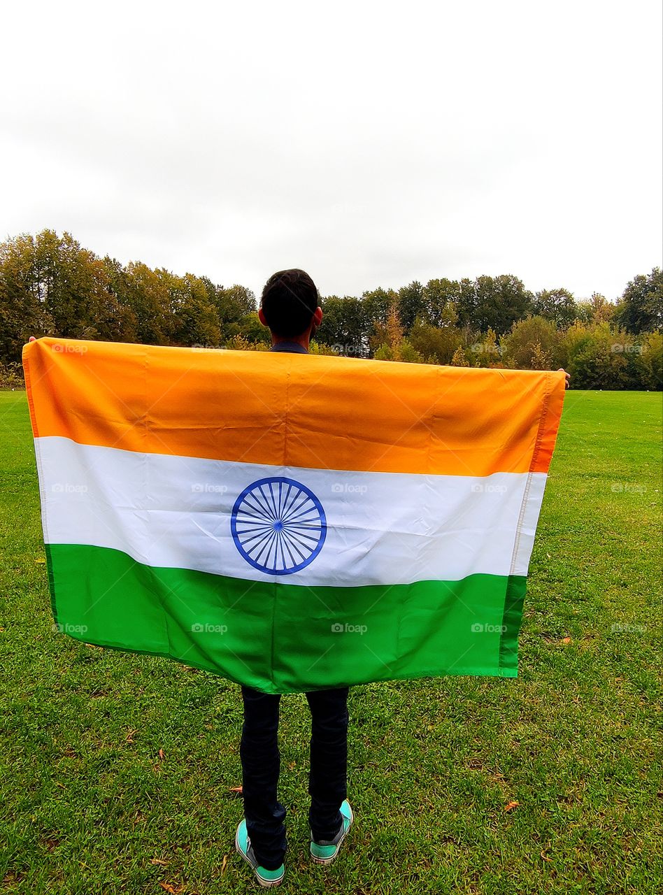 A man holds the flag of India.  Three horizontal stripes: saffron, white and green.  In the center of the flag is a 24-spoke, dark blue wheel