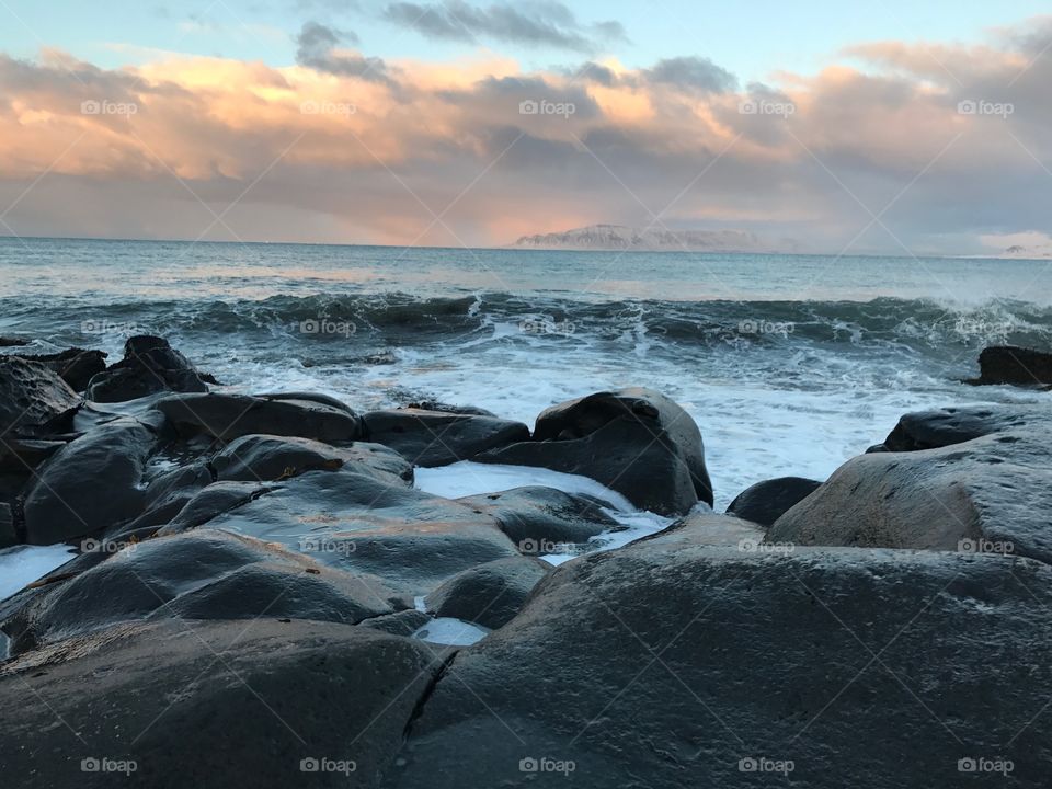 Quay in Reykjavik, wet black rocks on the Atlantic coast. A beautiful winter sunset with bright yellow and orange clouds against the background of the aquamarine sky. Waves and splashes.