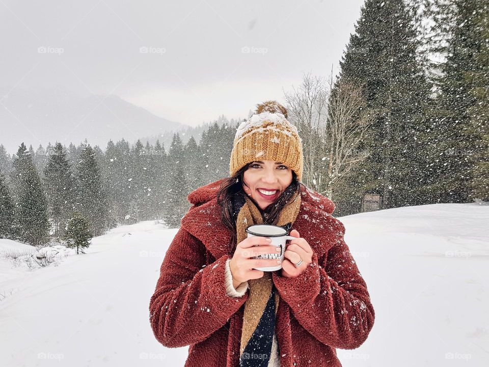 Winter portrait of young woman wearing winter clothes, holding cup of tea on meadow on a snowy day