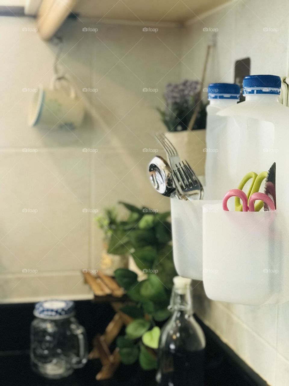 Plastic milk bottles converted into kitchen tools and holders . Here plast milk bottles contains some steel spoons forks and scissors. And surroundings covered with reusable glass containers and tea cup eco friendly flower pots.wooden stand