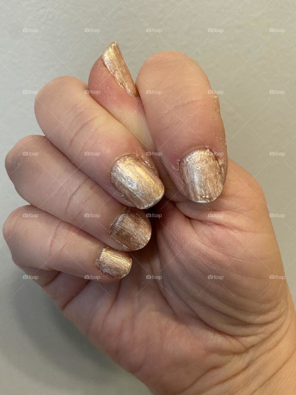 A hand in a fist with the thumb tucked in and peeking out, showing fingernails painted marbled-gold (a layer of white with a gold polish on top) against an off-white wall