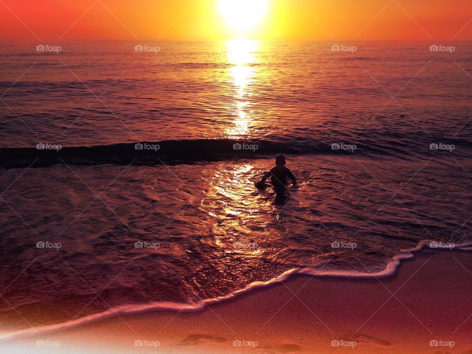 Small boy in the ocean surrounded by deep orange skies.