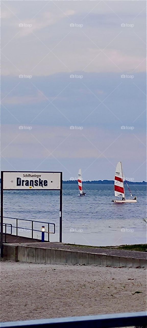 A sign on the dock by the “Baltic Sea” or “Ostsee” says that the town of “Dranske” is nearby on the island “Rügen” in “Mecklenburg - Western Pomerania”, Germany while sailboats with red and white stripes take to the wind. 2024. Hypnotic Productions