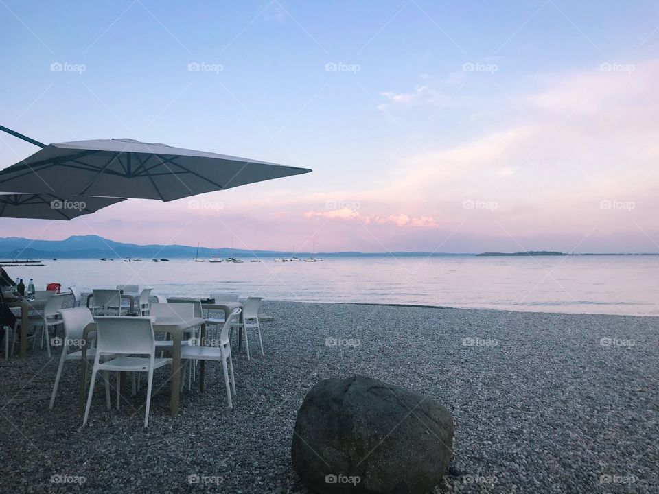 by colorful lake shore at sunset , there are many small stones, rocks , some sets of tables ,chairs. in the purple pink cloudy sky, and the color reflection on the lake surface. very romantic