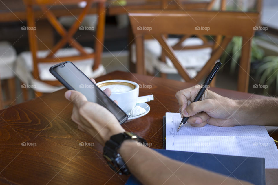 Man holds a smartphone in his hand and makes  the notes in notebook. Businessman working in cafe