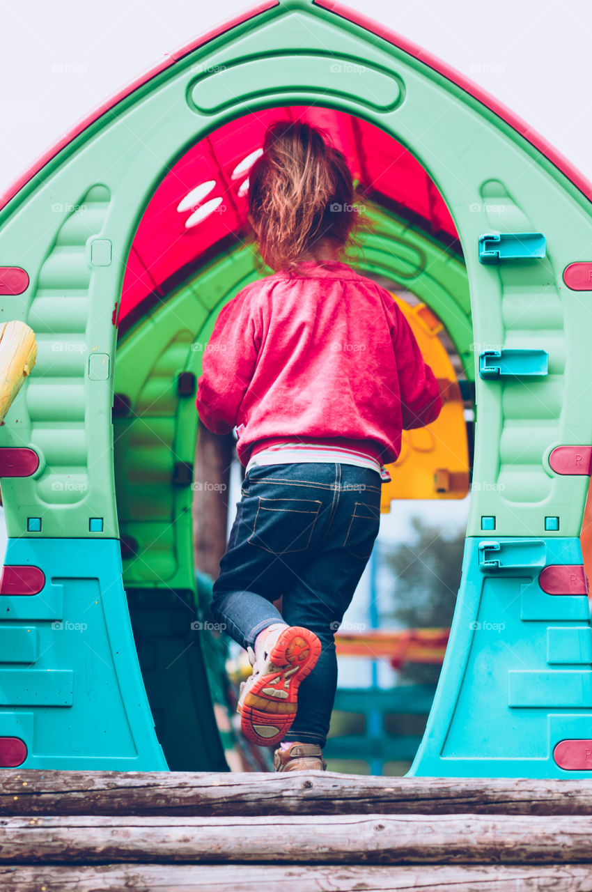 Little toddler girl playing in the playground going to plastic garden house on summer day