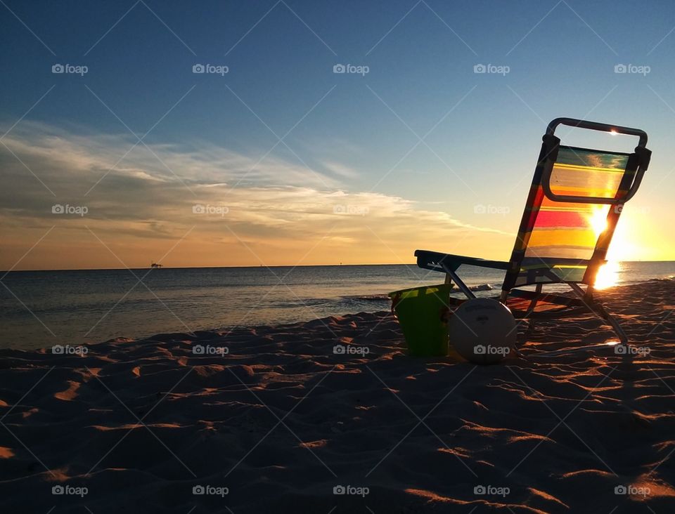 a beach chair, bucket, and volleyball all sitting on the sandy shore backlit by the gorgeous setting sun