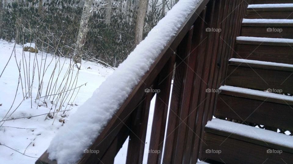Close-up of railing covered in snow with stairs on the right and bare trees and snow covered ground on the left.