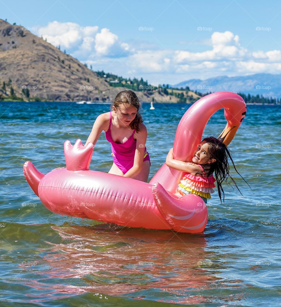Cousins playing on big pink flamingo toy on a scenic lake 