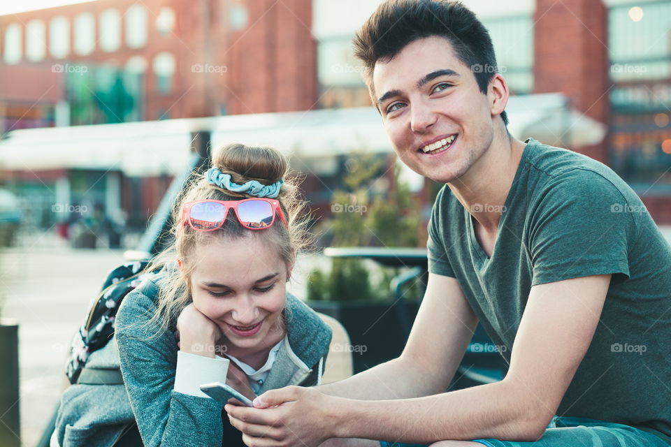 Couple of friends, teenage girl and boy, having fun with smartphones, sitting in center of town, spending time together