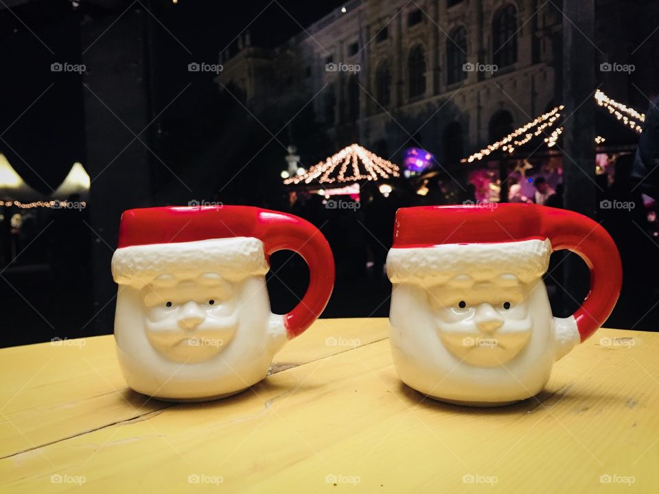 Two mugs shaped like Santa Claus on wooden table with Christmas lights in the background 