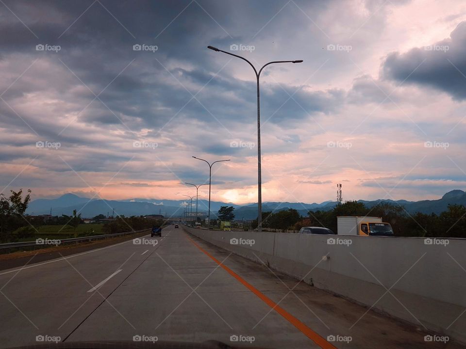 Driving on the Seroja toll road, Bandung city, on the edge toll road can see rice fields with green leaf tress, lighting poles on road medians, mountains and cloudy skies, yellowish red to gray in color.