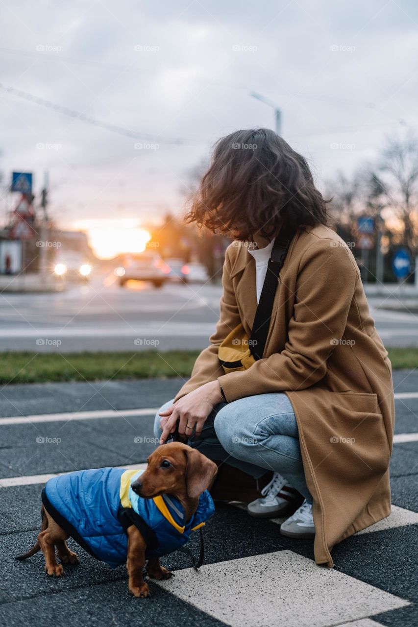 Millennial woman, dressed with a long coat, walking her small dog in the city, at sunset time, during winter.