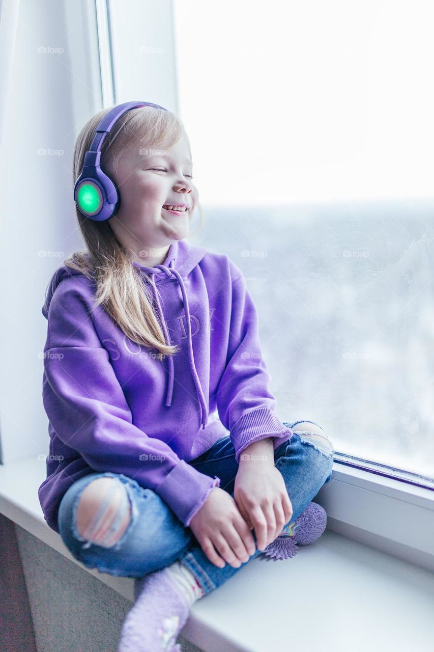 a girl in a purple sweatshirt sits by the window and listens to music through headphones