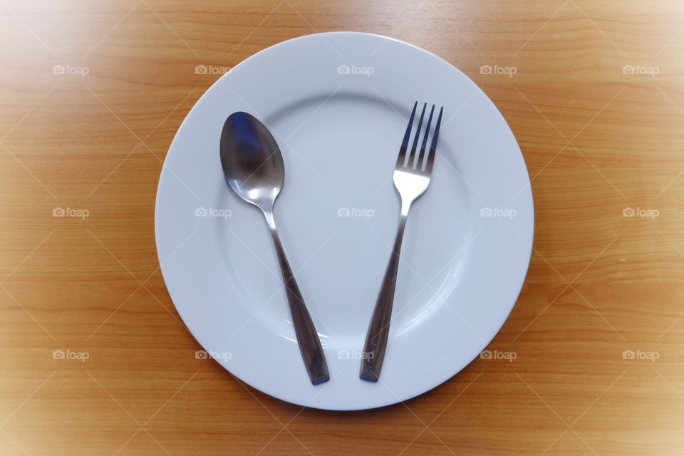 Empty white plate with spoon and fork on blown wooden background.