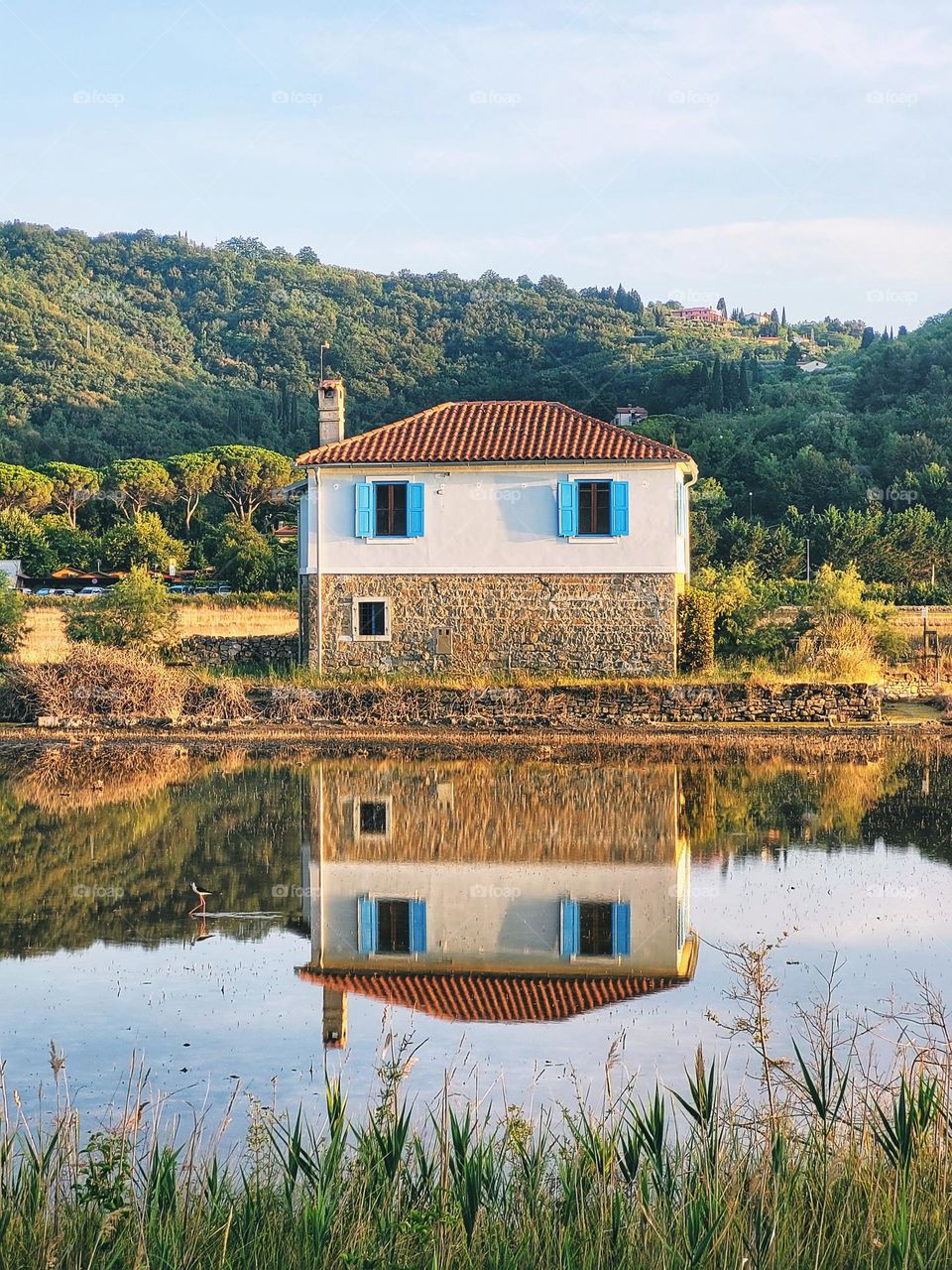 Scenic view of beautiful small house reflecting in the blue lake, river against blue sky and green forest.  Summer vacation.