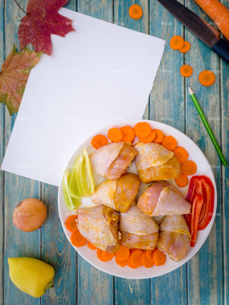 Pickled chicken legs with slices of carrots and green and red bell peppers on a white plate with a sheet of white paper for the recipe and a pencil on a wooden background.  Ingredients for the preparation of grilled chicken with vegetables.