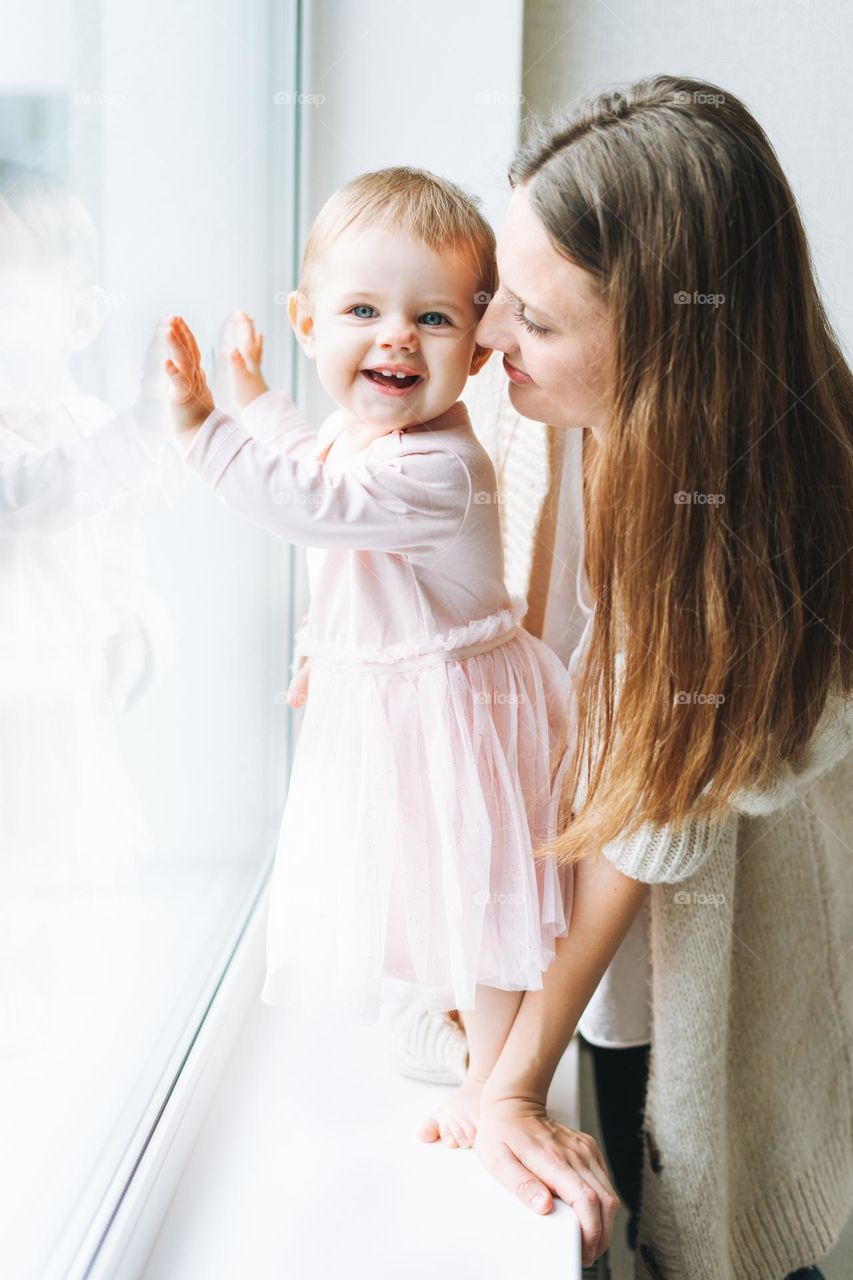 Young woman mom with long hair in cozy knitted cardigan with baby girl on hands near window at the home