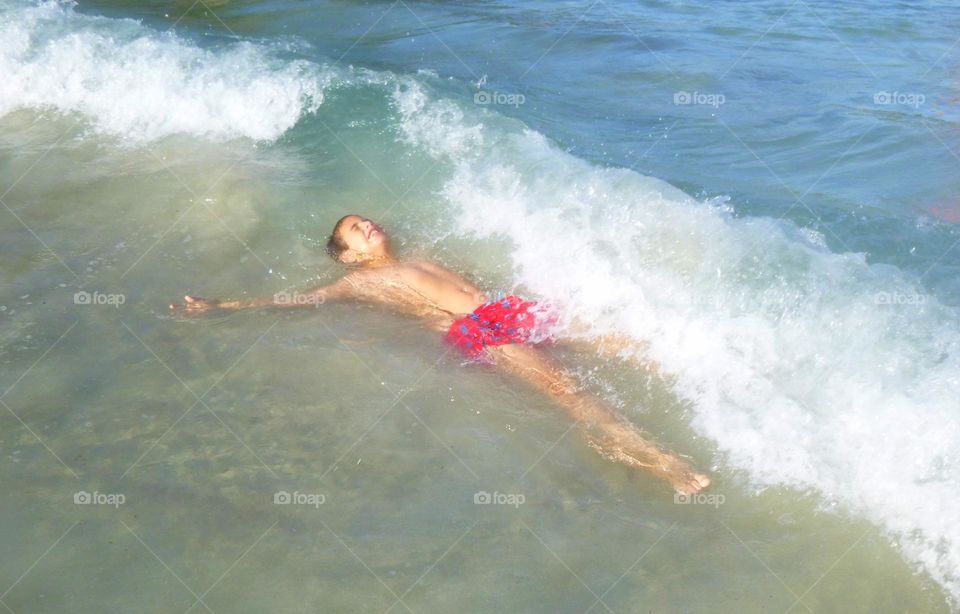 Boy in Red suimsuit, laughing, playing in the water at the sea, white a wave crashes against him