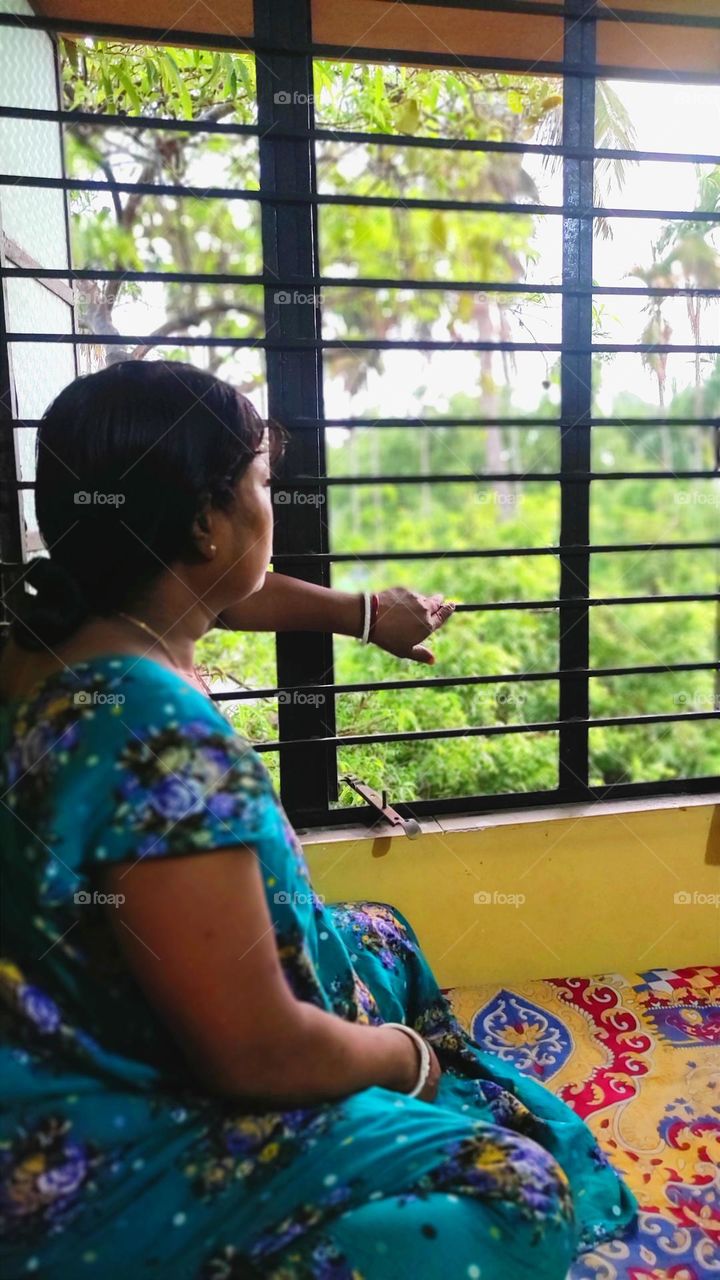 One Hindu Bengali Indian woman is watching beautiful trees through the window. She is sitting on a bed and holding one hand on the window.