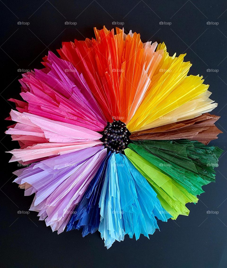 Flatlay composition of a multi-coloured flower made of silk papers and purple beads placed on black paper, creating a colour explosion