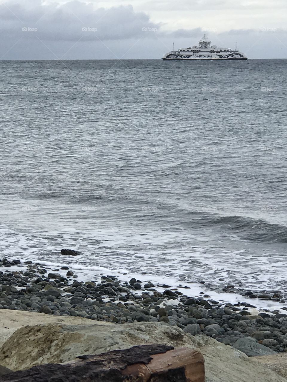 A shot of the ferry crossing the Strait on a grey, blustery day. The stony, rocky,  beach is in the foreground. The ferry is in the distance and behind it are grey and white clouds. 