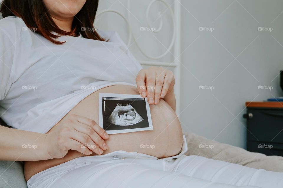 One caucasian young pregnant woman in white clothes holds a scanner photo of her baby while lying on the bed in the bedroom, close-up side view.