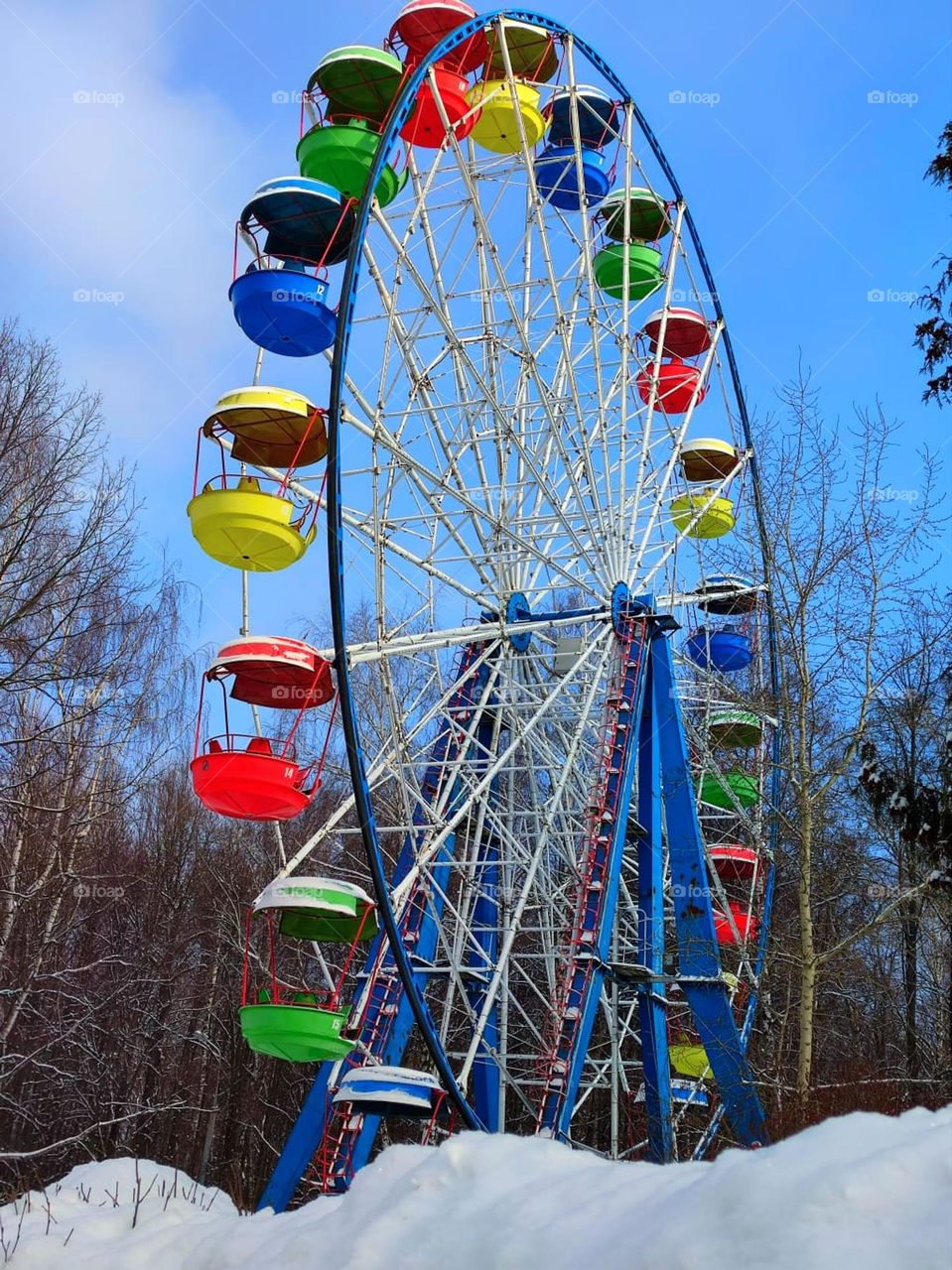 Winter park. Attraction "Ferris wheel" with multi-colored booths against the background of the blue sky and trees. In the foreground is a large white snowdrift