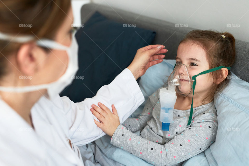 Doctor visiting little patient at home. Child having medical inhalation treatment with nebuliser. Woman wearing uniform and face mask