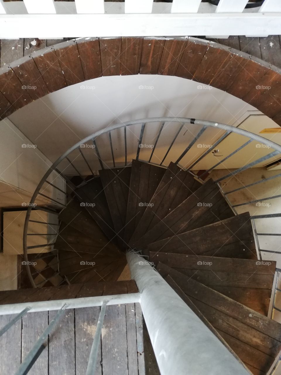 The brown much used spiral staircases leading down to the ground floor in an old building. The metallic railing is grey, the wall and the door painted white, the floor downstairs partly crisscross.